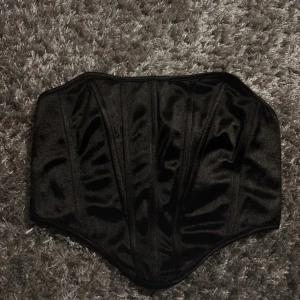 Svart satin korsett - Säljer en svart korsett i glansigt satinmaterial. Modellen är klassisk med markerade sömmar och formande passform. Perfekt att styla till olika outfits för en trendig look.