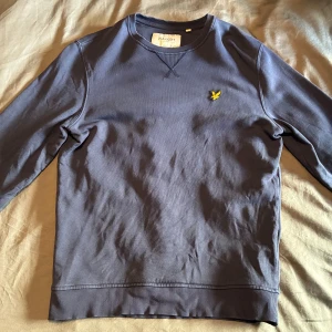 Blå sweatshirt från Lyle & Scott - Säljer en blå sweatshirt från Lyle & Scott med deras klassiska gula logga broderad på bröstet. Tröjan har rund halsringning och långa ärmar. Perfekt för en avslappnad stil.