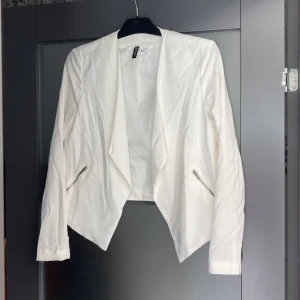 Vit kavaj med dragkedjedetaljer - Snygg vit kavaj från H&M med öppen front och dragkedjor vid fickorna. Jackan har lång ärm och en modern, asymmetrisk skärning framtill. Tillverkad i en blandning av polyester och viskos med lite elastan för stretch.
