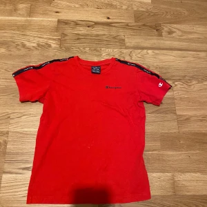 Röd t-shirt från Champion - Röd t-shirt från Champion med svarta och vita loggband längs axlarna. Klassisk passform och rund halsringning. Liten broderad Champion-logga på bröstet och ärmen. Perfekt för en sportig och avslappnad stil.