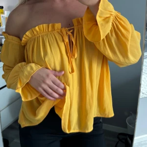 Gul offshoulder blus från NLY Trend - Säljer en solgul offshoulder blus från NLY Trend med volangdetaljer och knyt framtill. Blusen har långa, luftiga ärmar och en lös passform. Perfekt för att liva upp garderoben med lite färg!