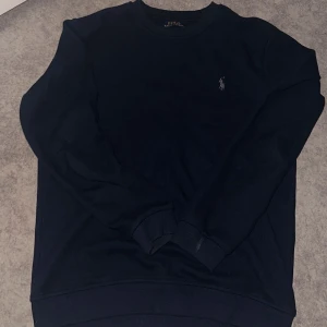 Mörkblå tröja från Polo Ralph Lauren - Mörkblå tröja från Polo Ralph Lauren med rund halsringning och diskret broderad logga på bröstet. Tröjan har långa ärmar och ribbade muddar vid ärmslut och nederkant. Perfekt för en stilren och klassisk look.