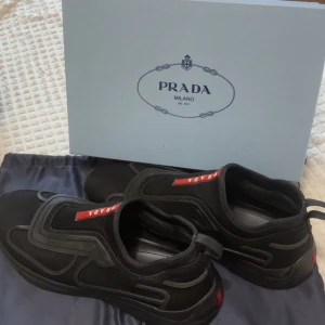 Svarta sneakers från Prada - Säljer ett par svarta sneakers från Prada med tydlig logga på plösen i rött och vitt. Skorna har en modern design med svarta detaljer och rund tå. Ovandelen är i textil och syntetmaterial. Perfekta för dig som gillar stilrena och exklusiva sneakers.