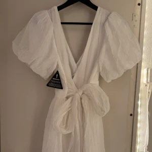 Studentklänning - the bow dress   - Superfin vit klänning med puffärmar. Klänningen har omlottdesign med knyt i midjan och är tillverkad i ett lätt, luftigt material. Perfekt för studenten!