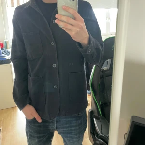 Svart overshirt från Uniqlo - Stilren svart overshirt från Uniqlo med tre knappar framtill och två stora fickor. Jackan är tillverkad i bomull med lite elastan för extra komfort. Perfekt för en avslappnad men ändå uppklädd look. Storleken är S men passar mer M.