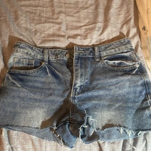 Blå jeansshorts med fransar - Säljer ett par klassiska blå jeansshorts med råa franskantade benslut och femficksmodell. Shortsen har bälteshällor och knappgylf. Perfekta för varma dagar och enkel att matcha med allt. Den är från Shein men det gör inte att  välja shein på varumärket 