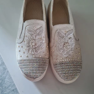 Vita slip-on sneakers med pärlor och blommor - Säljer ett par vita slip-on sneakers med platåsula. Skorna har broderade blommor, pärlor och glittriga stenar framtill samt längs sidorna. Perfekta för dig som vill sticka ut med detaljer och lite extra höjd.