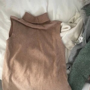 Beige stickad väst med polokrage - Säljer en beige stickad väst med hög polokrage. Västen är ärmlös och har en mjuk, ribbad struktur vid halsen. Perfekt att styla över en skjorta eller långärmad tröja.