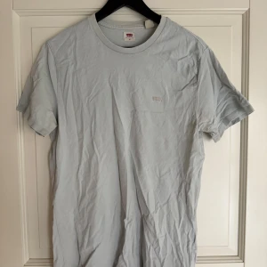 Ljusblå t-shirt från Levi's - Säljer en stilren ljusgrå t-shirt från Levi's med diskret broderad logga på bröstet. Klassisk passform och rund halsringning. Perfekt till jeans eller shorts för en clean look.
