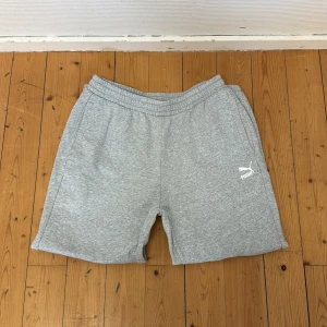 Gråa mjukisbyxor från Puma - Snygga grå mjukisbyxor från Puma med elastisk midja och snörning. Tillverkade i mjuk bomullsmix och har två sidofickor. Perfekta för träning eller chill. Puma-logga i vitt på vänster ben.