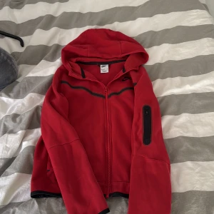 Röd Nike tech hoodiejacka med dragkedja - Röd hoodiejacka med hel dragkedja framtill och svarta detaljer. Jackan har huva och en praktisk ficka med dragkedja på ärmen. Perfekt för en avslappnad stil eller till träning.
