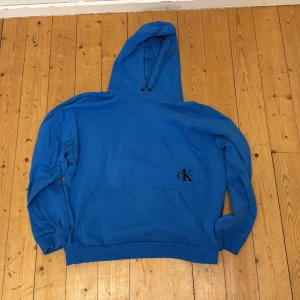 Blå hoodie från Calvin Klein - Snygg blå hoodie från Calvin Klein med svart CK one-logga broderad på framsidan. Klassisk känguruficka och huva med dragsko. Perfekt för en avslappnad och stilren look.