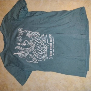 Grön t-shirt med tryck från H&M - Säljer en grön t-shirt från H&M med vitt tryck framtill som föreställer en bläckfisk och texten 'Captain's Wharf'. T-shirten har rund hals och korta ärmar. Perfekt för en avslappnad stil.