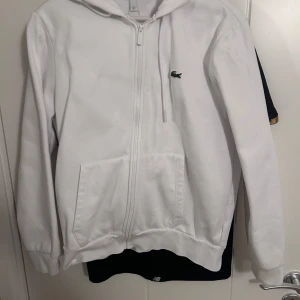 Vit hoodie med dragkedja från Lacoste - Säljer en vit hoodie från Lacoste med dragkedja framtill och klassisk krokodillogga på bröstet. Tröjan har huva, två fickor och ribbade muddar vid ärmslut och nederkant. Perfekt för en avslappnad stil.