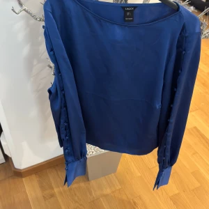 Blå blus med knappar från Lindex - Säljer en blå blus från Lindex med dekorativa knappar längs ärmarna och knyt vid ärmsluten. Blusen har lång ärm och är tillverkad i ett glansigt, mjukt material som ger en elegant känsla.