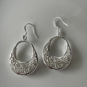Silver 925 örhängen med blommönster - Säljer ett par snygga silvriga örhängen med öppet ovalt mittparti och detaljerat blommönster. Perfekta för att ge en extra touch till din stil. Lätta att bära och passar till många tillfällen.