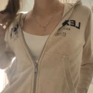 Beige hoodie från Lexington - Säljer en beige hoodie från Lexington med dragkedja och huva. Tröjan har blå logotyptryck framtill och fickor på magen. Perfekt för en avslappnad stil och passar året runt.