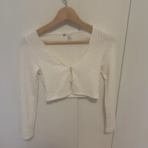 Vit croppad ribbad topp med knyt - Säljer en vit croppad topp med lång ärm och v-ringning. Toppen har ett ribbat mönster och knyt framtill, vilket ger en trendig look. Perfekt att styla med jeans eller kjol.