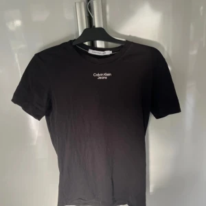 Svart t-shirt från Calvin Klein Jeans - Säljer en svart t-shirt från Calvin Klein Jeans med liten vit logga tryckt på bröstet. Klassisk rund halsringning och korta ärmar. Tillverkad i 100% bomull för en skön och mjuk känsla.