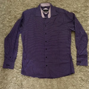 Lila rutig skjorta från Allan Clark - Säljer en snygg lila skjorta från Allan Clark med diskret rutigt mönster och svarta knappar. Skjortan har lång ärm och ljuslila insida på kragen. Perfekt för dig som vill ha en stilren look.