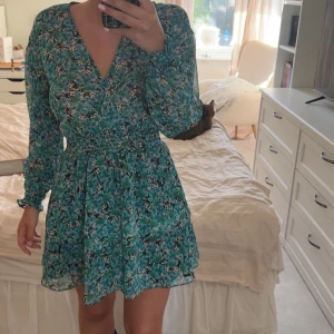 Blommig grön omlottklänning - Säljer en söt grön kortklänning med blommigt mönster och omlott-design. Klänningen har långärmade ärmar, markerad midja och volangdetaljer nertill. Perfekt för vår och sommar!