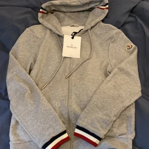 Grå hoodie från Moncler med dragkedja - Säljer en grå hoodie från Moncler med dragkedja och huva. Jackan har ribbade muddar med röd, vit och marinblå rand, samt Moncler-logga på ärmen. Snygga metalländar på snörena och klassisk sportig look.