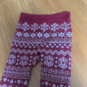 Vinröda leggings med snöflingor och hjärtan - Mysiga vinröda leggings med vita snöflingor, hjärtan och zigzag-mönster. Perfekta för vintersäsongen och jul, med bred resår i midjan för extra komfort. Mjukt och stretchigt material som passar bra till kalla dagar. Dom har 2 hål i rumpan