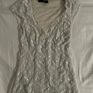 Vit spets topp med v-ringning - Söt vit topp med v-ringad hals och spetsdetaljer över hela plagget samt bar rygg. Tröjan har korta ärmar och en liten rosett framtill. Perfekt för somriga tillfällen eller studentfirande.