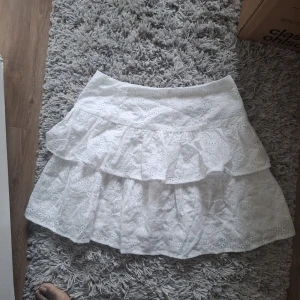 Vit volangkjol med broderade detaljer - Säljer en söt vit kjol med volanger och broderade blommönster. Kjolen har en kort längd och är perfekt för varma dagar. Den är tillverkad i bomull och har en luftig och feminin känsla.