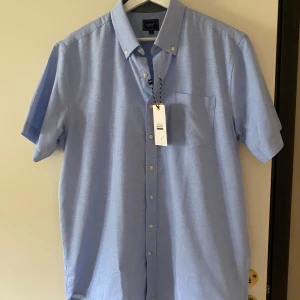 Ljusblå kortärmad skjorta från Signal - Säljer en ljusblå kortärmad skjorta från Signal Red Label i storlek L. Skjortan har klassisk krage, bröstficka och knappar framtill. Perfekt för varmare dagar och enkel att matcha med olika outfits. Helt ny!!