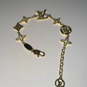 Guldigt armband med kristaller från Luis Vitton  - Exklusivt armband i guldton med LV-logga och ikoniska LV-blommor dekorerade med gnistrande kristaller. Justerbar kedja med LV-berlock. Perfekt för dig som vill ha något lyxigt och trendigt.