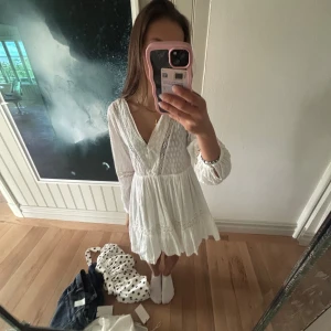 Vit spetsklänning med v-ringning - Söt vit klänning med v-ringning och spetsdetaljer. Långärmad modell med lätt volang nertill och markerad midja. Perfekt för sommarens alla tillfällen.