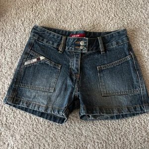 Lågmidjade jeansshorts  - Snygga mörkblå jeansshorts med två stora framfickor och två bakfickor. Shortsen har dubbla knappar i midjan och synliga sömmar i kontrastfärg. Perfekta för sommaren och har en klassisk jeanslook.