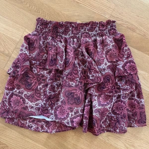 Rosa mönstrad kortkjol med volang - Supersöt kortkjol i rosa och vinröda toner med paisleymönster och volangdetaljer. Kjolen har resår i midjan för extra komfort och är fodrad med vitt tyg på insidan. Vet ej storlek men skulle säga M/L.