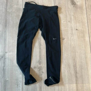 Svarta träningstights från Nike - Svarta långa träningstights från Nike med liten logga på benet. Tightsen har en tight passform och dragkedja vid benslutet. Perfekta för löpning eller gym.