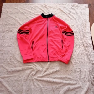 Rosa adidas tränings jacka. - Säljer en rosa Adidas träningsjacka med svarta detaljer och dragkedja framtill. Jackan har svarta ränder på ärmarna och svart krage. Perfekt för dig som gillar sportig stil och vill sticka ut med färg.