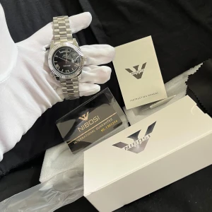 Silverfärgad klocka från Nibosi med svart urtavla - Stilren klocka från Nibosi med silverfärgad länk i metall och svart urtavla. Klockan har tydliga index, datumvisning och dagvisning. Levereras med originalförpackning, garanti och instruktionsmanual.