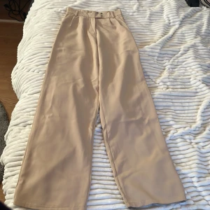 Beiga vida kostymbyxor från Outfitbook - Säljer ett par beiga kostymbyxor från Outfitbook med hög midja och vida ben. De har blivit insydda i midjan och en bit ner för en bättre passform men som går att sprätta upp om man skulle vilja. Säljs för 90kr+frakt 