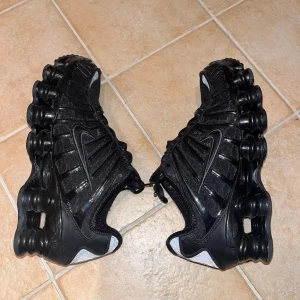Svarta Nike Shox sneakers - Säljer ett par svarta Nike Shox sneakers med tydliga Shox-dämpningar i sulan och reflekterande detaljer baktill. Skorna har snörning, mesh- och syntetpaneler samt röda Nike Shox-loggor på plös och häl.