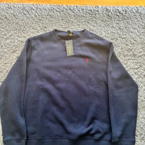 Mörkblå sweatshirt från Polo Ralph Lauren - Säljer en klassisk mörkblå sweatshirt från Polo Ralph Lauren med rund halsringning och den ikoniska röda loggan broderad på bröstet. Tröjan har långa ärmar och är tillverkad i mjuk bomull, perfekt för en stilren och avslappnad look.