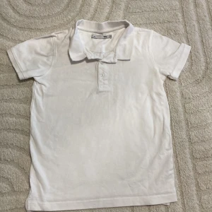 Vit pikétröja i bomull - Klassisk vit pikétröja med krage och två knappar framtill. Tröjan är tillverkad i mjuk bomull och har korta ärmar. Perfekt för en stilren och enkel look.