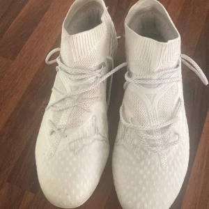 Vita fotbollsskor med strumpliknande skaft - Säljer ett par vita fotbollsskor från Puma med strumpliknande skaft och snörning. Skorna har en mönstrad ovandel i syntetmaterial och fasta dobbar för spel på gräs. Perfekta för dig som vill ha stilrena och lätta skor på planen.