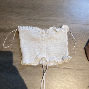 Vit smockad tubtopp från Bik Bok - Säljer en söt vit smockad tubtopp från Bik Bok med volangdetaljer upptill och nedtill samt knytband framtill. Perfekt för varma dagar och enkel att matcha med jeans eller kjol.