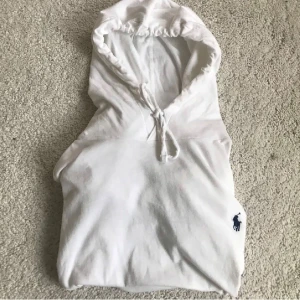 Vit Ralph Lauren hoddie - Säljer en klassisk vit hoodie från Polo Ralph Lauren med dragsko i huvan och den ikoniska blå loggan broderad på bröstet. Perfekt för en clean och stilren look.