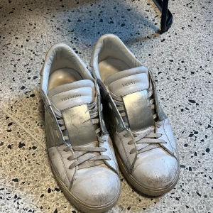 Vita Valentino Open sneakers med silverdetalj - Säljer ett par vita Valentino Open sneakers med bred silverfärgad rem över vristen. Skorna har klassisk snörning och rund tå. Snygg och stilren design med diskret Valentino-logga på plösen. Använda men ändå i ett rätt okej skick 6,5/10 Kan tänka mig gå ner i pris vid snabb affär!