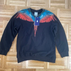 Svart sweatshirt med färgglada vingar från Marcelo Burlon - Cool svart sweatshirt från Marcelo Burlon med ikoniskt vingtryck i blått och rött över axlar och bröst. Tröjan har rund halsringning och ribbade muddar vid ärmslut och nederkant. Perfekt statement piece för din garderob.