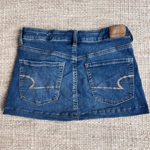 Blå jeanskjol från American Eagle - Säljer en klassisk blå jeanskjol från American Eagle med fem fickor och snygga kontrastsömmar. Kjolen har två bakfickor med dekorativa sömmar och en liten läderpatch i midjan. Perfekt till sneakers eller boots!