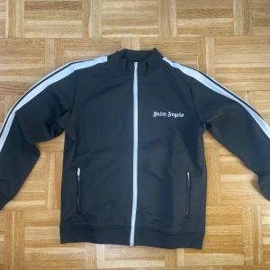 Palm Angels kofta - Palm Angels svart track jacket med vita ränder längs ärmarna och vit dragkedja framtill. Jackan har ståkrage, två fickor med dragkedja och logga på bröstet. Perfekt för en sportig och trendig stil.
