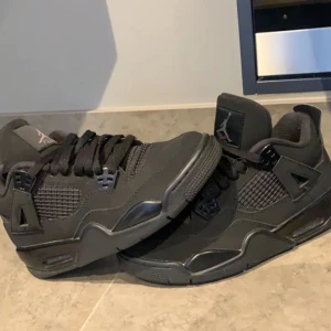 Svarta  Jordan 4 black cat - Säljer ett par helsvarta  Jordan 4 Retro med klassisk mesh och skinnpaneler. Skorna har rund tå, platt sula och ikoniska detaljer som synliga plastvingar och Jumpman-logga på plösen. Perfekta för dig som gillar streetwear och sneakerskultur.
