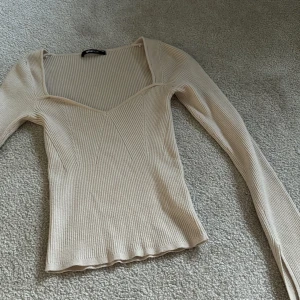 Beige ribbad topp från Gina Tricot - Säljer en beige ribbad topp från Gina Tricot i storlek XS. Toppen har lång ärm, v-ringad halsringning och figurnära passform. Perfekt att styla till olika outfits!